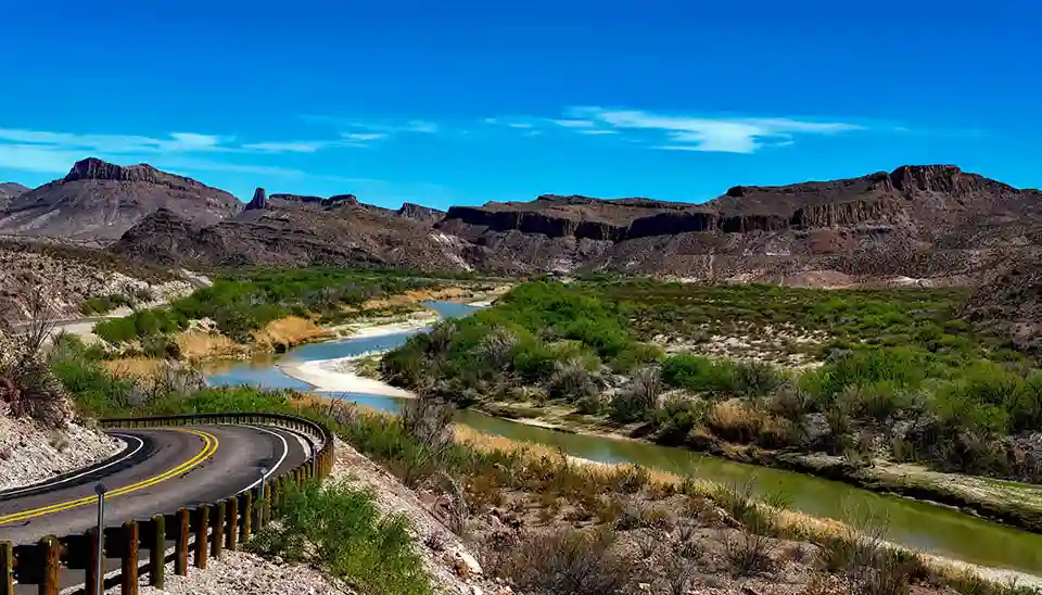 Texas landscape featuring the Rio Grande River flowing through arid land with the only lush plants snuggled up to the bank. Rocky hills and formations are in the background set against a blue sky with faint clouds that gradually gets lighter as it drops behind the rock formations.